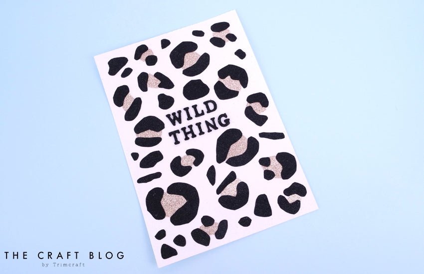 A white card features bold black and gold leopard spots made with the Dovecraft Glitter Card A4 Pad Classic by Printable Heaven, "WILD THING" in the center, on a light blue background. "THE CRAFT BLOG by Trimcraft" is printed at the bottom left.