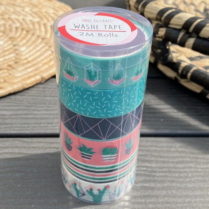 A stack of Printable Heaven Love to Craft Washi Tape Tube 6 Rolls - Cactus (393872), with geometric and potted plant designs, sits on a gray surface beside woven mats. The label reads Washi Tape 2M Rolls.