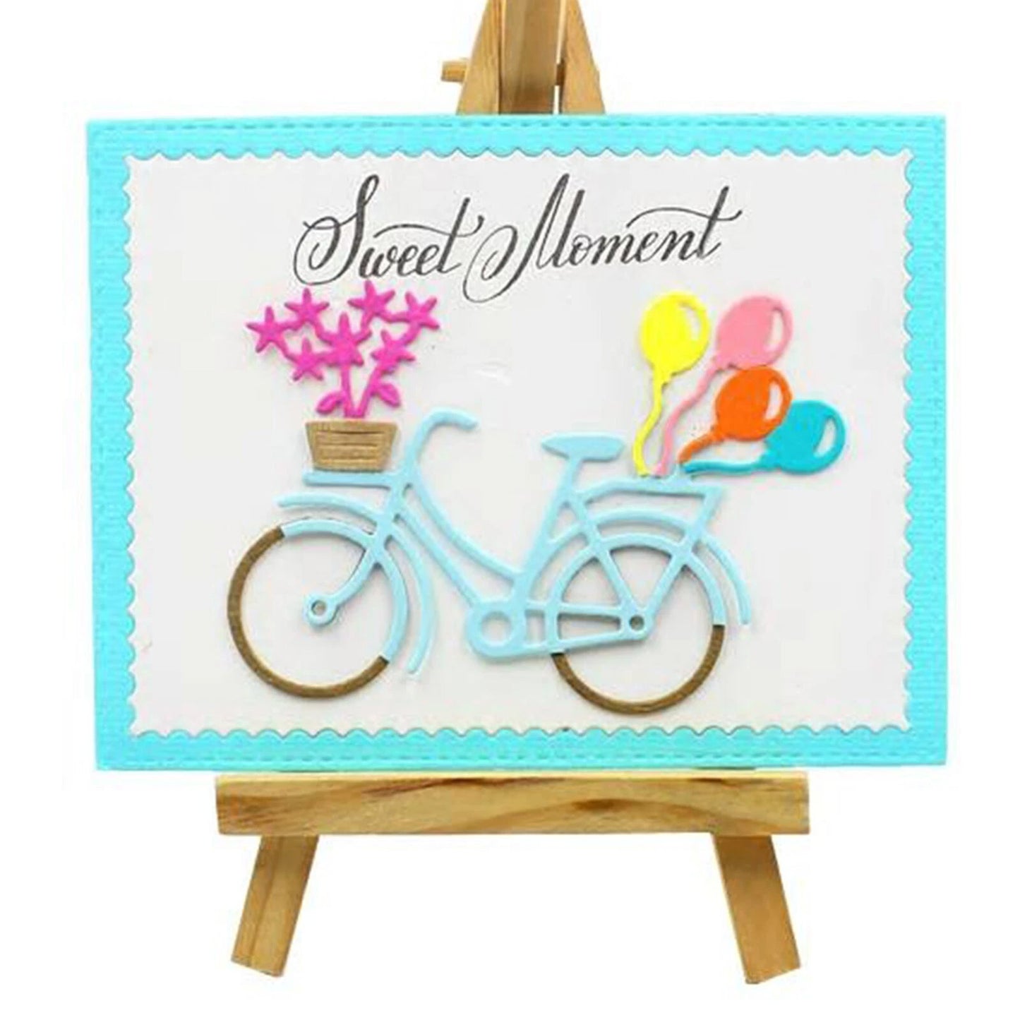 A handmade card on an easel, featuring a blue bicycle created with the Printable Heaven Bicycle with Balloons & Gifts Cutting Die (D104). The basket is adorned with pink flowers and colorful balloons, and the design is accented with charming paper crafting embellishments. The words “Sweet Moment” are written above.