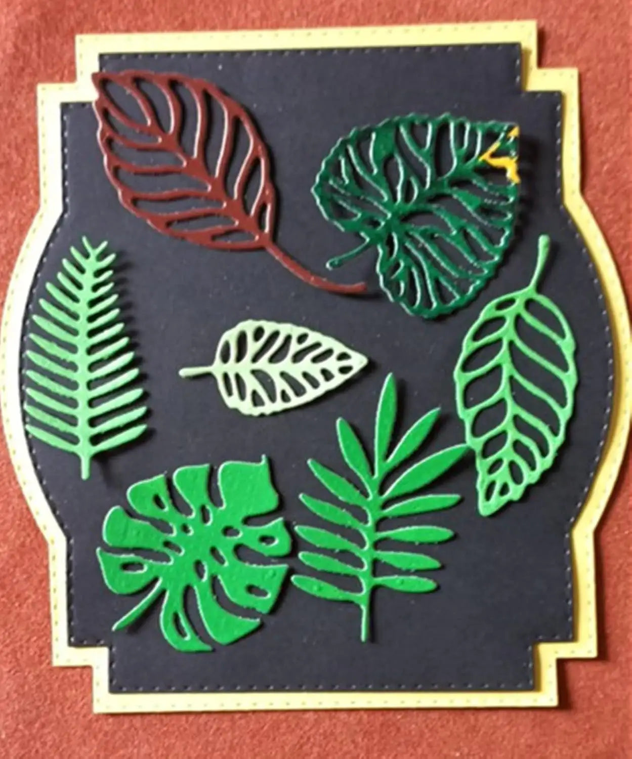 A handmade card featuring seven intricate, cut-out leaves in green and brown, arranged on a black background with a yellow border—perfect for card making and botanical paper crafts using your favorite Tropical Leaves Cutting Die.