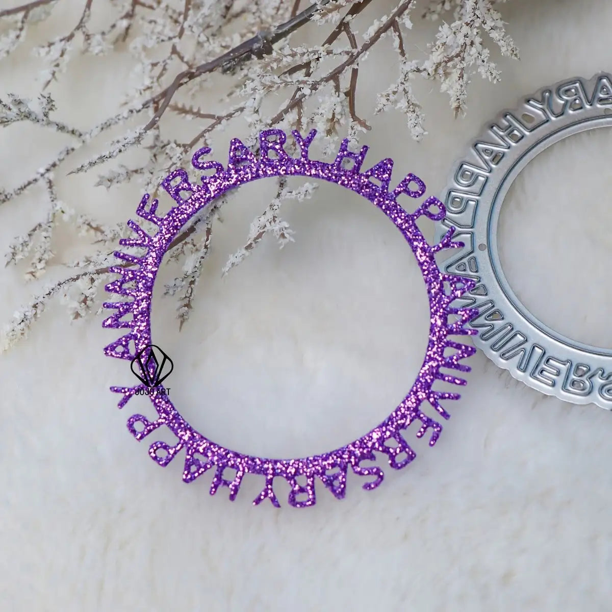 A purple glittery circular piece, created using the Printable Heaven Happy Anniversary Cutting Die (1 PC) (W151), displays the words “HAPPY ANNIVERSARY” and is shown resting on a white furry surface, paired elegantly with frosted branches and part of a silver ornament nearby.