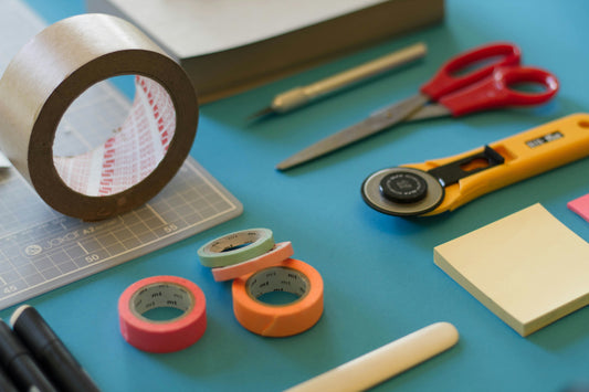 A collection of crafting tools including a penknife, scissors, sticky note pads and masking tape.