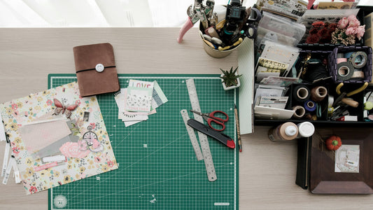 A crafting table with various tools on it, including a box of sewing accessories, scissors, rulers and cards.