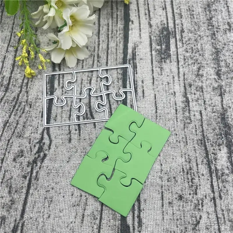 The Printable Heaven Jigsaw Puzzle Cutting Die (6 PCS) (T071) is shown alongside green puzzle pieces on a textured surface, with white flowers in the background—ideal for cardmaking or scrapbooking projects.