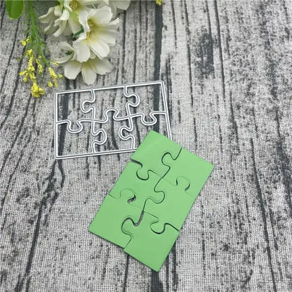 The Printable Heaven Jigsaw Puzzle Cutting Die (6 PCS) (T071) is shown alongside green puzzle pieces on a textured surface, with white flowers in the background—ideal for cardmaking or scrapbooking projects.