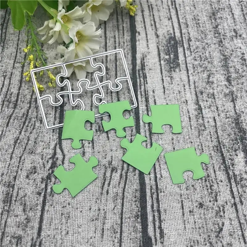 Six green pieces from the Printable Heaven Jigsaw Puzzle Cutting Die (6 PCS) (T071) and a sturdy metal puzzle die frame rest on a textured gray surface, accented by delicate white flowers in the background—an ideal scene for your cardmaking or scrapbooking projects.