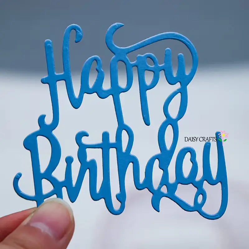 A hand holds a blue cutout made with the Happy Birthday Sentiment Cutting Die (1 PC) (W243) by Printable Heaven, featuring “Happy Birthday” written in cursive letters. The background is blurred, and the words DAISY CRAFTS alongside a small flower appear on the right, making it perfect for birthday cards or scrapbooking projects.