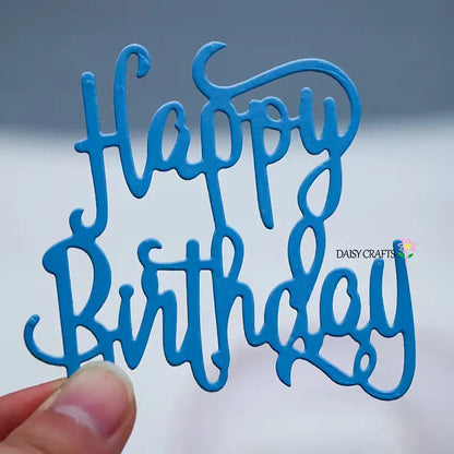 A hand holds a blue cutout made with the Happy Birthday Sentiment Cutting Die (1 PC) (W243) by Printable Heaven, featuring “Happy Birthday” written in cursive letters. The background is blurred, and the words DAISY CRAFTS alongside a small flower appear on the right, making it perfect for birthday cards or scrapbooking projects.