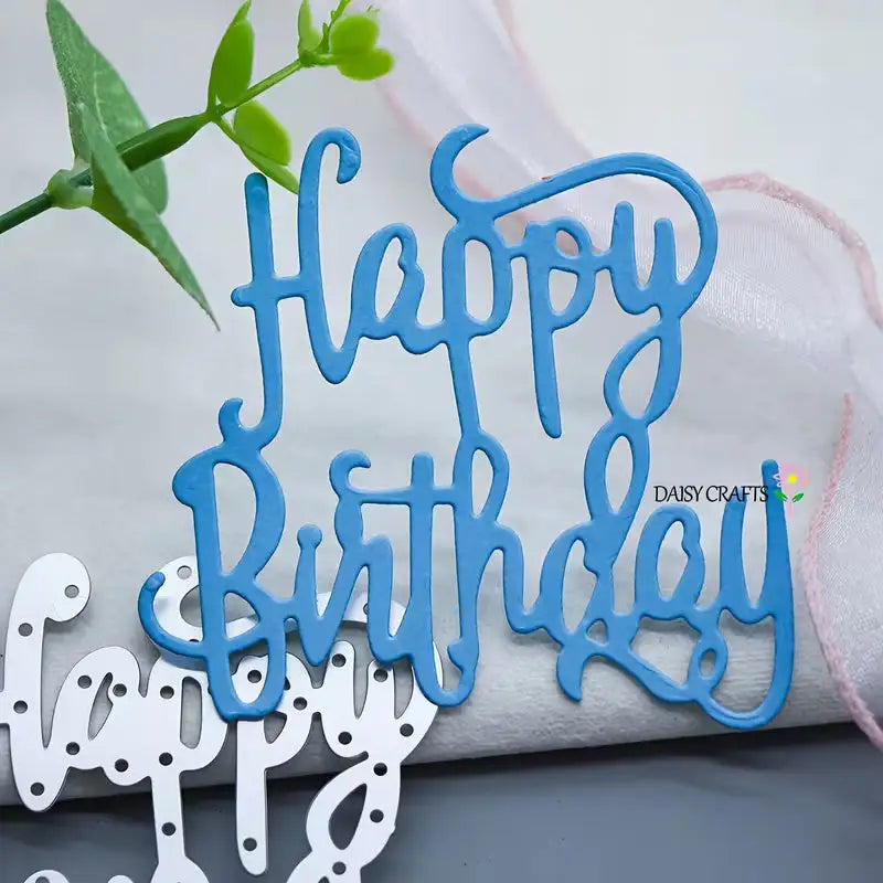 A blue “Happy Birthday” cutout, crafted with the Printable Heaven Happy Birthday Sentiment Cutting Die (1 PC) (W243), is featured with a green plant and white fabric ribbon in the background—perfect for scrapbooking or making one-of-a-kind birthday cards. Below, a white version of the “Happy Birthday” sentiment made using the same cutting die lies flat.