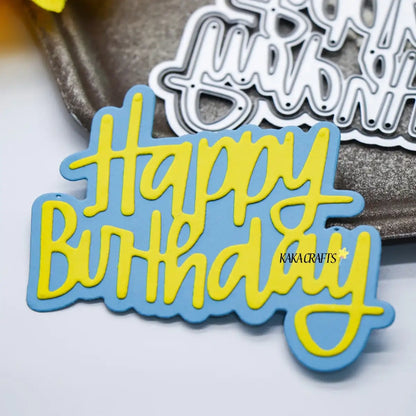 A blue and yellow Happy Birthday cutout card, created with the Printable Heaven Happy Birthday Sentiment Cutting Die (2 PCS) (W101), sits on a metallic surface—perfect for paper-crafting projects—with a matching die and part of a yellow ribbon visible in the background.