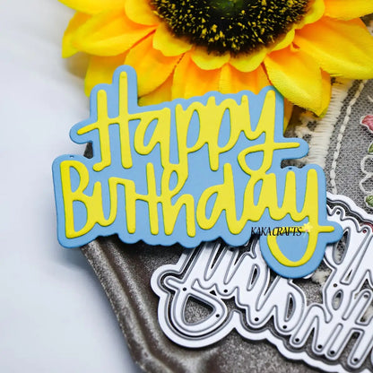 A yellow and blue “Happy Birthday” sign rests beside a large artificial sunflower and metal dies from the Printable Heaven Happy Birthday Sentiment Cutting Die (2 PCS) (W101) set, displayed on a textured surface—perfect inspiration for paper-crafting projects like creating birthday cards with your favorite Printable Heaven die.