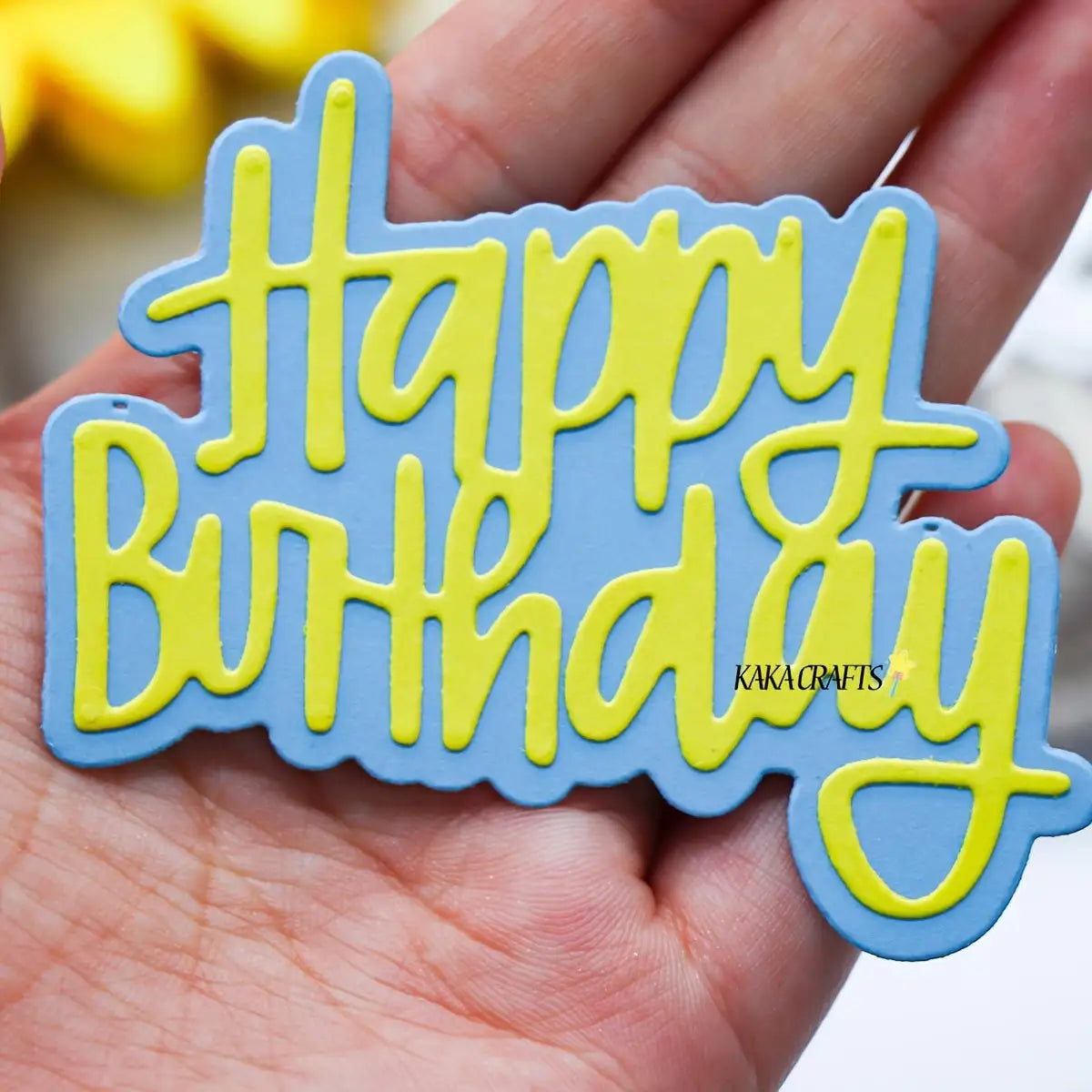 A hand displays a blue and yellow “Happy Birthday” sign crafted with layered material and elegant cursive lettering, perfect for paper-crafting projects or birthday card making using the Printable Heaven Happy Birthday Sentiment Cutting Die (2 PCS) (W101).