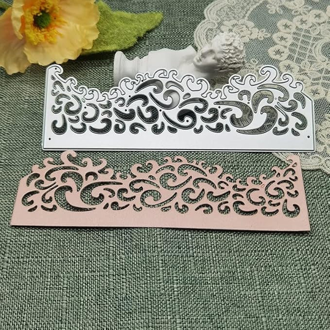 Showcased on a green textured surface with a flower, lace doily, and white decorative bust are two intricately cut rectangles—one metallic, one pink—demonstrating the delicate detailing possible with the Printable Heaven Ornate Filigree Border Cutting Die (1 PC) (B004). Their swirling wave patterns highlight the die’s suitability for elegant papercraft projects.