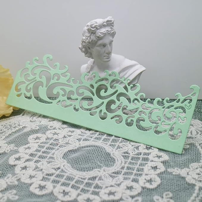 An Ornate Filigree Border Cutting Die (1 PC) (B004) from Printable Heaven, featuring a decorative green lace-patterned design, is displayed on a lace tablecloth with a small white classical bust in the background.