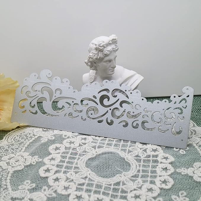 An Ornate Filigree Border Cutting Die (1 PC) (B004) by Printable Heaven, featuring intricate swirly cutout patterns reminiscent of decorative silver filigree, is displayed on a lace cloth with a small white bust sculpture in the background.