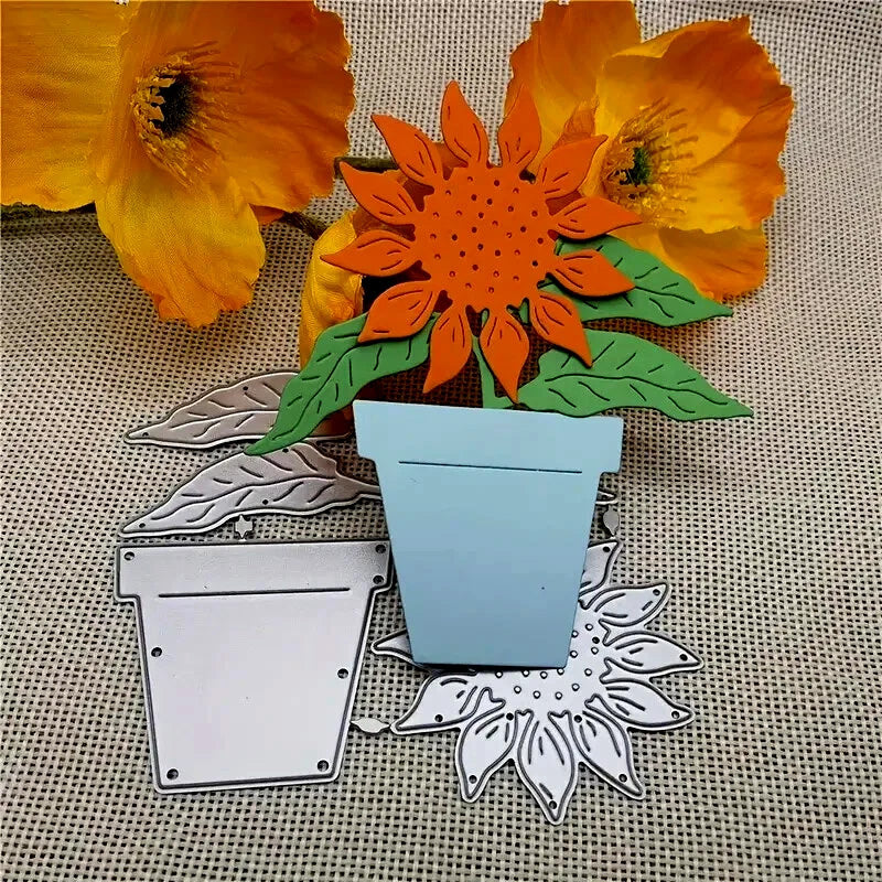 Paper cutouts of a sunflower in a blue pot are displayed, featuring intricate botanical details made with the Printable Heaven Sunflower & Pot Plant Cutting Die (3 PCS) (F036). Additional metal cutting dies and large orange flowers are arranged on a burlap surface in the background, creating an inspiring scene for creative paper crafting.