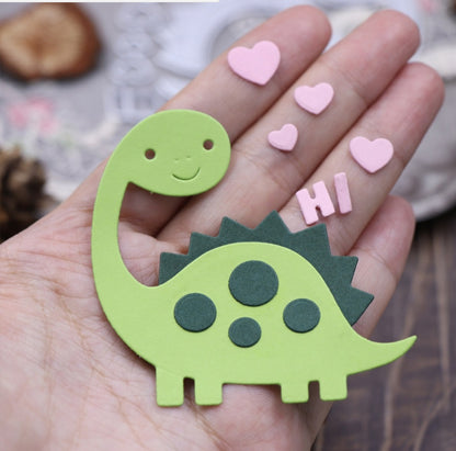 A hand displays an adorable green cartoon dinosaur cutout, crafted using the Printable Heaven Cute Dinosaur Card Cutting Die – Layered Craft Die Set (11 PCS) (A212). The smiling dinosaur, accented with dark green spots, is surrounded by pink hearts and “HI” letters—perfect embellishments for card making or scrapbooking projects.