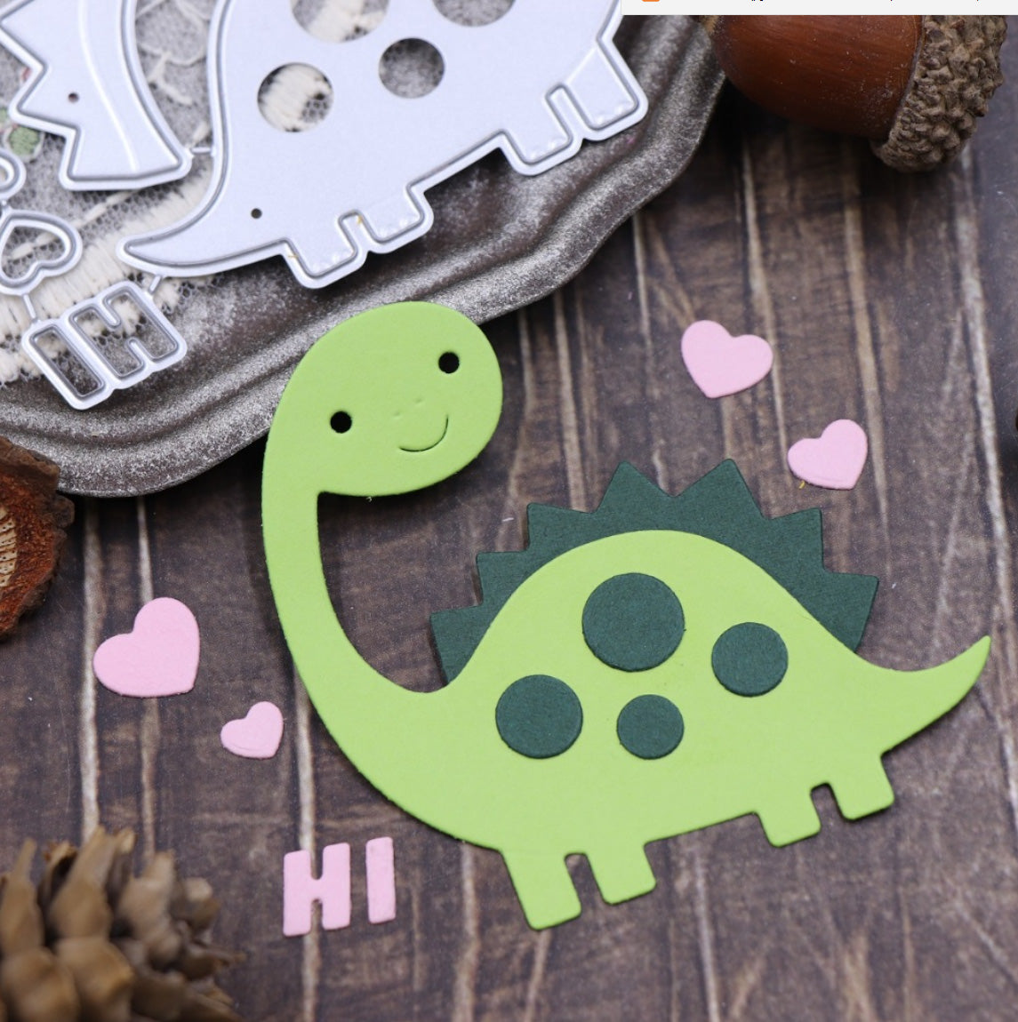 A charming green paper dinosaur with dark green spots and spikes, made using the Printable Heaven Cute Dinosaur Card Cutting Die – Layered Craft Die Set (11 PCS) (A212), is surrounded by pink hearts and the word “HI” on a wooden surface. The metal cutting dies—ideal for scrapbooking or card making—and some acorns are placed nearby.