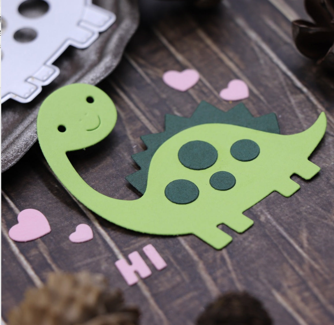 The Cute Dinosaur Card Cutting Die – Layered Craft Die Set (11 PCS) (A212) from Printable Heaven features an adorable green dinosaur with a friendly smile, pink hearts, and the word “HI,” perfect for adding charm to your scrapbooking or card making projects.