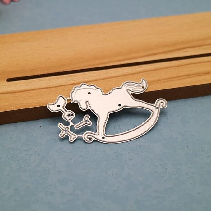 A Printable Heaven Rocking Horse Cutting Die set (4 pcs) (A108), ideal for use with die-cutting machines, is displayed on a blue surface beside a wooden piece featuring a long slot.