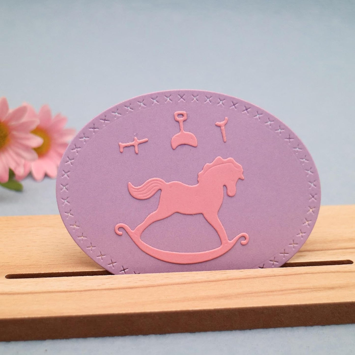 Showcasing a handmade oval card with a stitched border in purple, this design uses the Printable Heaven Rocking Horse Cutting Die (4 pcs) (A108) and die-cutting machines to feature a pink rocking horse and baby toys. Displayed on a wooden stand, it’s ideal as baby shower décor, with a touch of pink flower visible on the left.