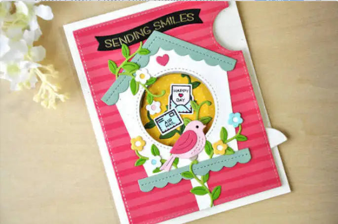 Crafted with the Printable Heaven Large Cuckoo Clock Cutting Die (13 PCS) (A030), this handmade greeting card showcases a delightful chalet-inspired birdhouse, a pink bird, flowers, and leaves. The card features “SENDING SMILES” and “HAPPY DAY” sentiments on a pink striped background, finished with delicate white flowers in the top left corner.