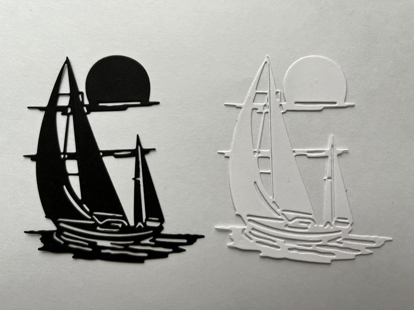 Two sailboat silhouettes, one black and one white, appear side by side—each sailing on water with a sun or moon above, made from solid and outlined shapes on a plain background. This simple design is perfect inspiration for nautical crafts or for using the Printable Heaven Sailing boat/Yacht on the Water Cutting Die (1 PC) (D098) in your creative projects.