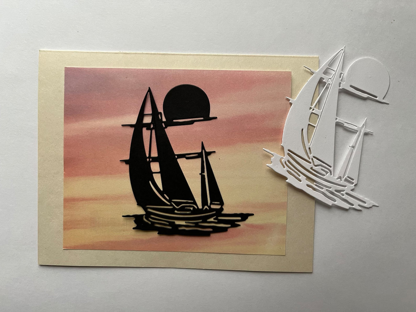 A card featuring a sunset and a black sailboat silhouette is displayed next to a white sailboat cutout, both beautifully crafted with the Printable Heaven Sailing boat/Yacht on the Water Cutting Die (D098), on a white surface.