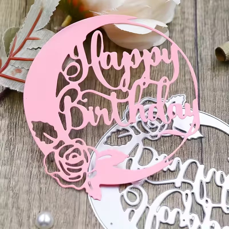 A pink paper cutout showcases a “Happy Birthday” sentiment inside a circle adorned with a rose at the bottom, displayed on a wooden surface next to the Printable Heaven Happy Birthday Sentiment Cutting Die (1 PC) (DJ022) and decorative flowers—ideal for birthday card making and other creative papercraft projects.