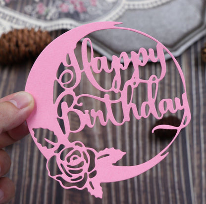 A hand displays the Printable Heaven Happy Birthday Sentiment Cutting Die (1 PC) (DJ022)—a delicate papercraft piece featuring a cursive “Happy Birthday” with a rose motif, shown atop a wooden table—perfect inspiration for birthday card making and creative DIY projects.