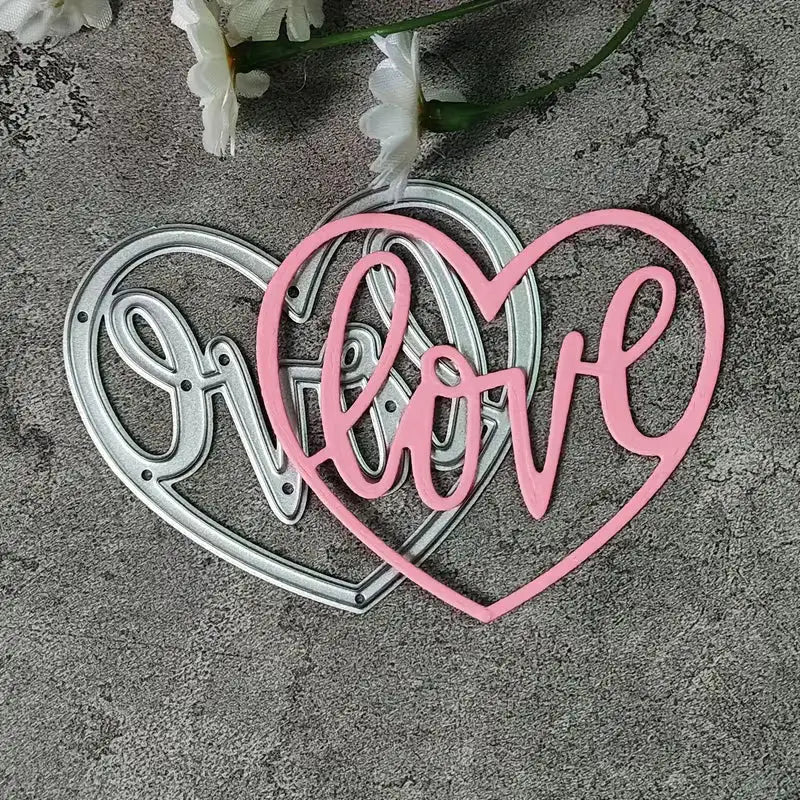 A silver and a pink “Love Heart Cutting Die” (W094) by Printable Heaven are showcased on a gray textured background. The pink die features the word “love” in elegant cursive, evoking a delicate, romantic touch ideal for wedding crafts. White flowers appear at the top of the image, completing this inspirational scene.