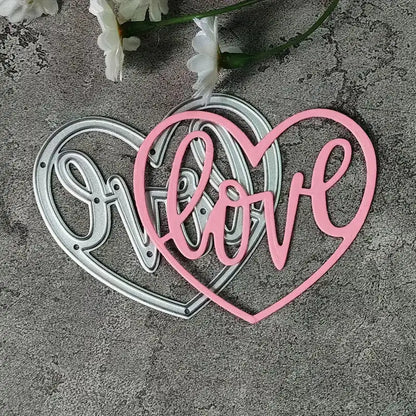 A silver and a pink “Love Heart Cutting Die” (W094) by Printable Heaven are showcased on a gray textured background. The pink die features the word “love” in elegant cursive, evoking a delicate, romantic touch ideal for wedding crafts. White flowers appear at the top of the image, completing this inspirational scene.