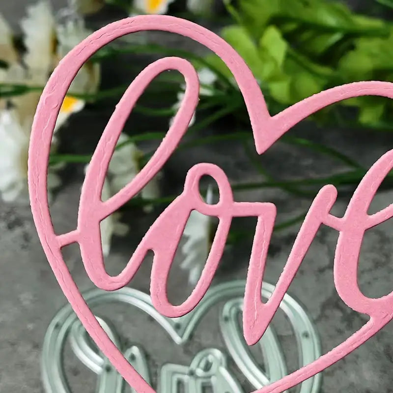 Crafted with the Love Heart Cutting Die (1 PC) (W094) from Printable Heaven, this pink heart-shaped decoration showcases the word “love” in graceful cursive and is photographed against a softly blurred backdrop of green leaves and white flowers—ideal for wedding crafts or romantic occasions.