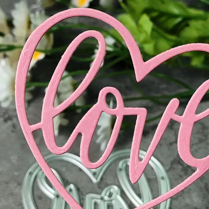 Crafted with the Love Heart Cutting Die (1 PC) (W094) from Printable Heaven, this pink heart-shaped decoration showcases the word “love” in graceful cursive and is photographed against a softly blurred backdrop of green leaves and white flowers—ideal for wedding crafts or romantic occasions.