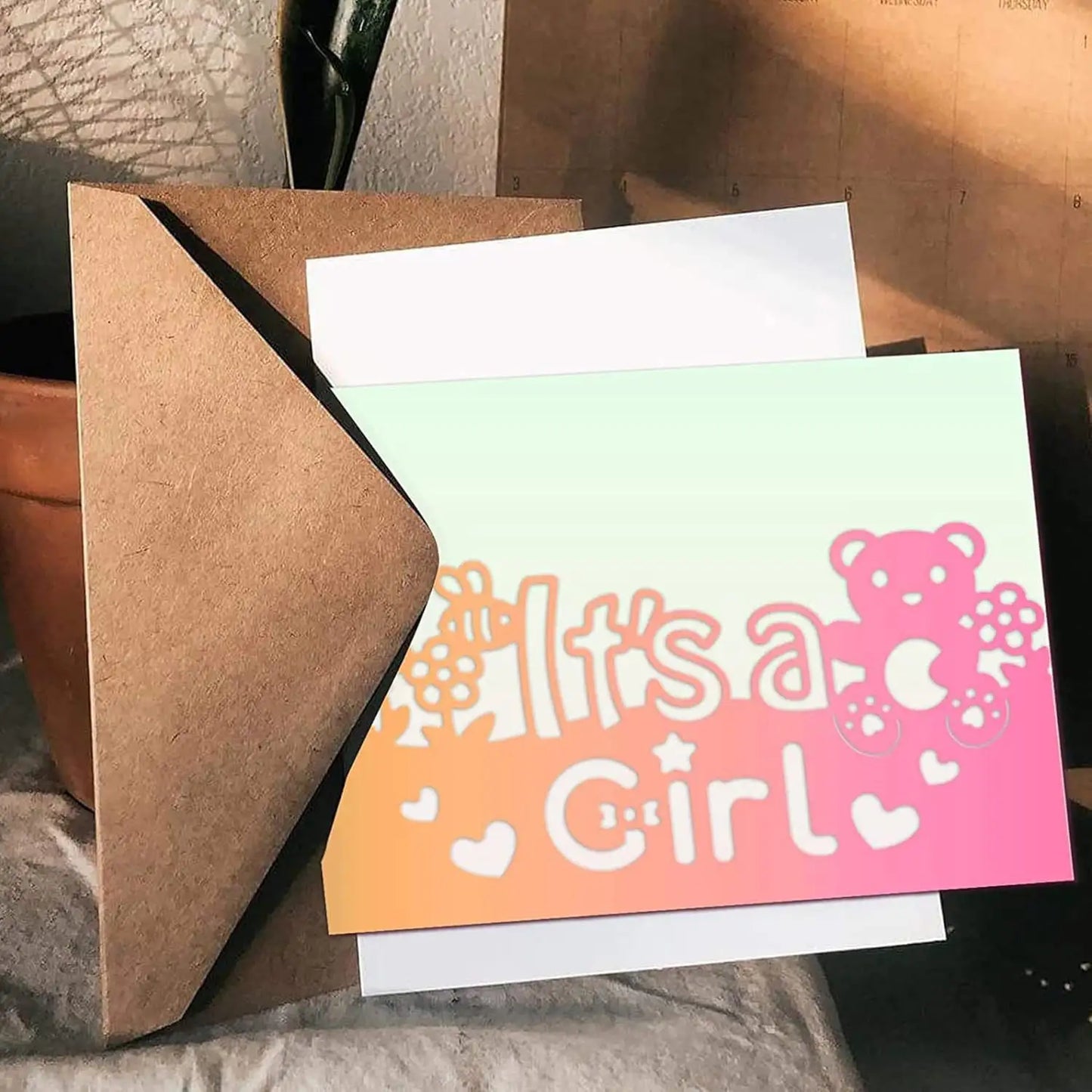 A greeting card featuring an “It’s a Girl” message in a pink and orange gradient, adorned with hearts, flowers, and a teddy bear—created using the Printable Heaven It’s a Girl Sentiment Cutting Die (1 PC) (W082)—is partially tucked inside a brown envelope on the table, making it an ideal addition to your baby shower decorations.