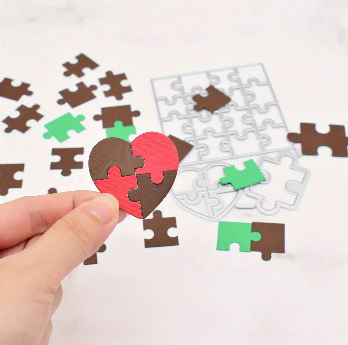 A hand holds a heart-shaped puzzle made of red and brown pieces, expertly crafted using the Printable Heaven Jigsaw Puzzle Cutting Die (21 PCS) (T204). Additional puzzle pieces, including green ones, are scattered on a white surface next to an incomplete puzzle outline—ideal for paper crafts or designing personalised puzzles.