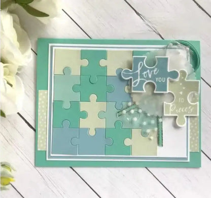 A charming handmade card featuring pastel puzzle pieces, crafted with the Printable Heaven Jigsaw Puzzle Cutting Die (21 PCS) (T204) and adorned with the heartfelt message “Love you to pieces,” rests on a white wooden surface beside delicate white flowers—a delightful addition to any paper crafts collection.