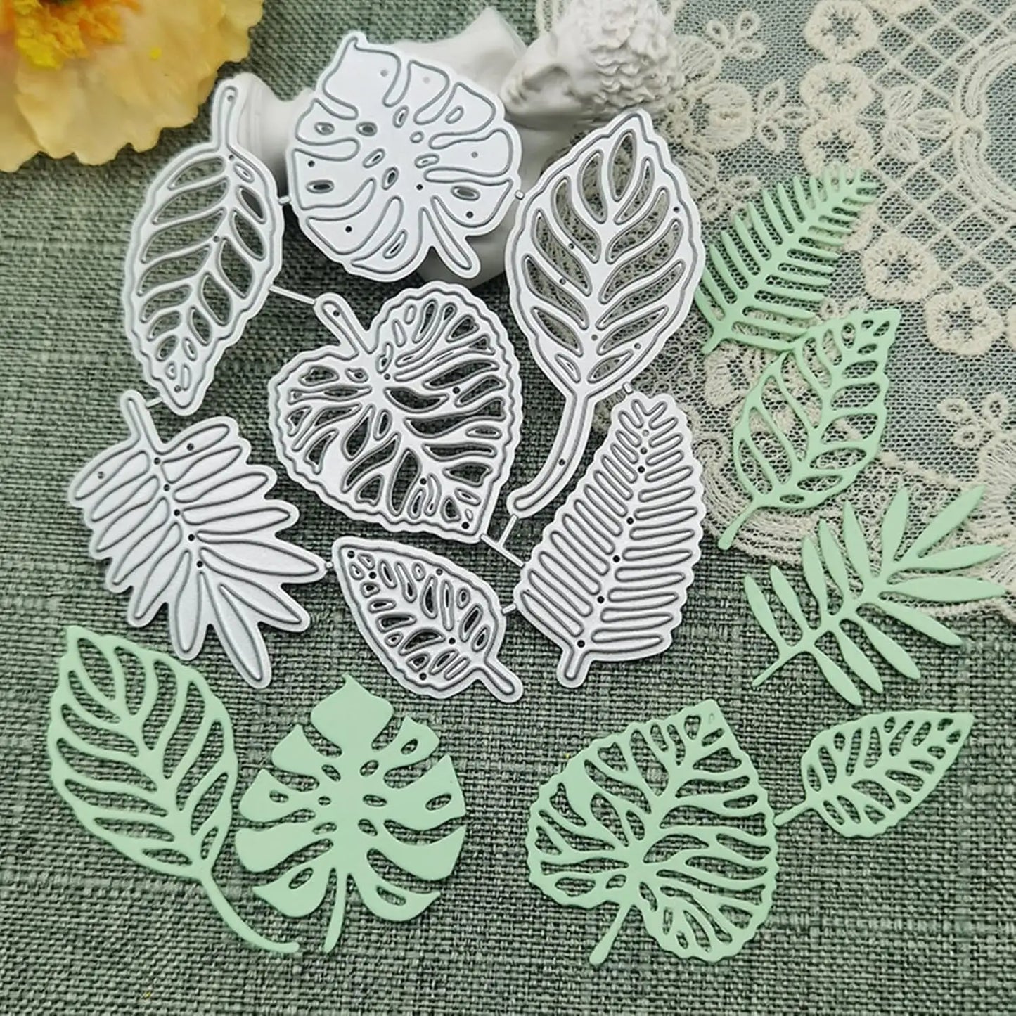 Printable Heaven’s Tropical Leaves Cutting Die Set (7 PCS) (L011) and their delicate light green paper cutouts are beautifully arranged on a textured green fabric, accented by lace and a yellow flower in the background. These metal cutting dies are perfect for botanical paper crafts and card making, adding a lush, natural touch to your creative projects.