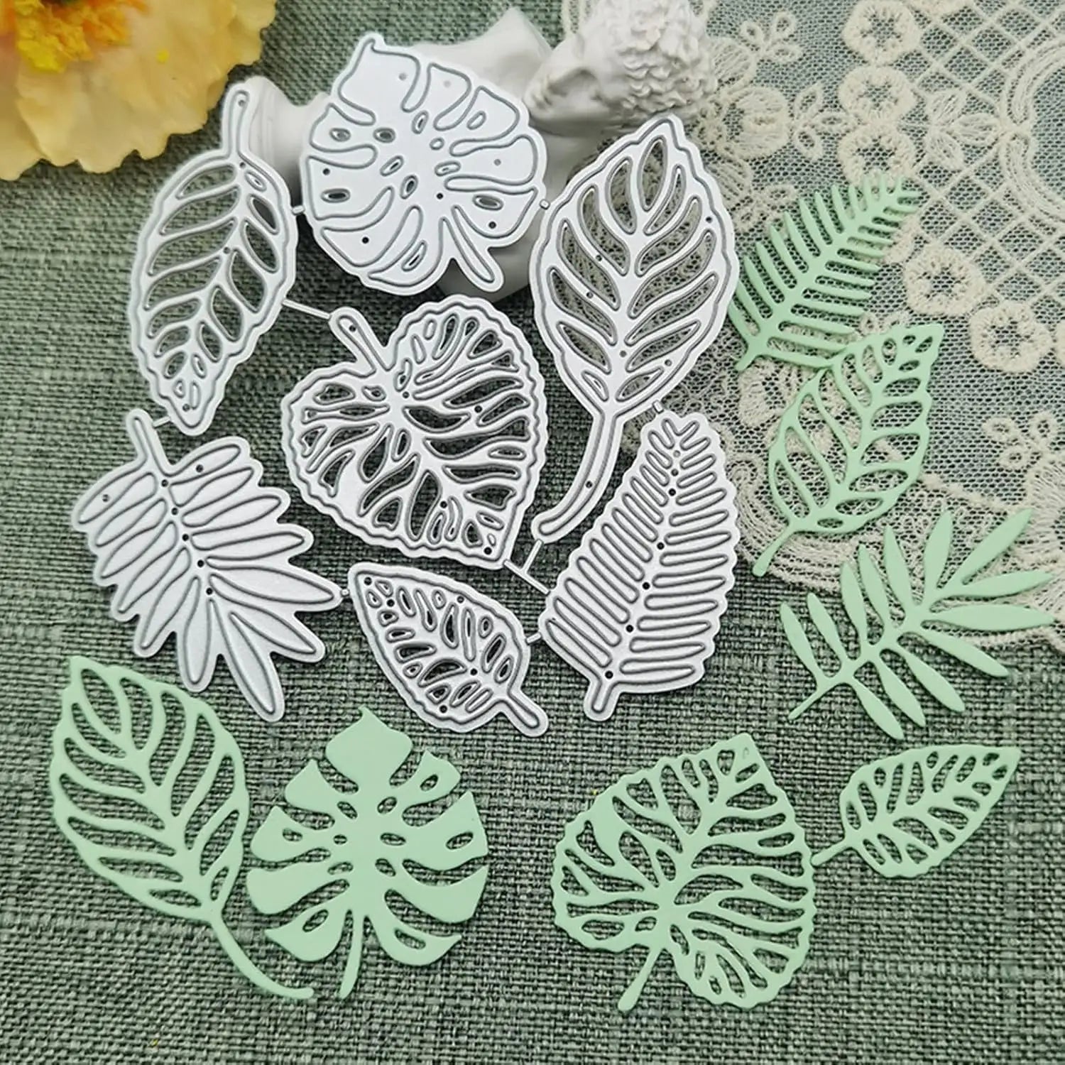 Printable Heaven’s Tropical Leaves Cutting Die Set (7 PCS) (L011) and their delicate light green paper cutouts are beautifully arranged on a textured green fabric, accented by lace and a yellow flower in the background. These metal cutting dies are perfect for botanical paper crafts and card making, adding a lush, natural touch to your creative projects.