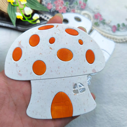 A hand holds the Printable Heaven Cutting Die – Playful Mushroom House Design (3 PCS) (L002), featuring a charming, cartoon-style mushroom home with an orange door, window, and spots—ideal for woodland or fairy-tale themed crafts. In the background are a tray with yellow flowers and a floral mat.