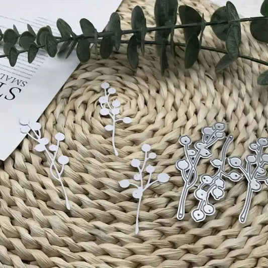 Three metal cutouts from the Printable Heaven Christmas Berries & Nature Cutting Die Set (3 PCS) (DJ075)—featuring branch and round berry designs—are displayed on a woven mat beside a green leafy twig and a piece of white paper, making them ideal for nature-inspired crafts.
