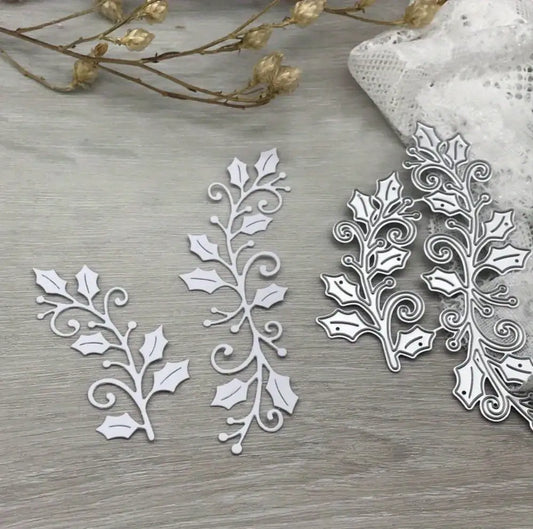 Two Printable Heaven Christmas Holly Cutting Dies (2 PCS) (SP109) and their delicate white paper cutouts, shaped like festive holly branches, rest on a wooden surface—ideal for adding a touch of elegance to your Christmas crafting. Dried flowers and lace accent the corner, inspiring creative papercraft projects.