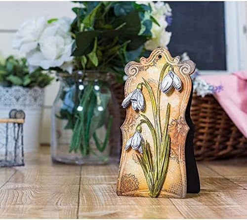 The Large Snowdrop Cutting Die (1 PC) (F147) by Printable Heaven is displayed upright on a wooden floor, showcasing elegant raised white snowdrop designs—perfect inspiration for handmade cards or other papercrafting projects. In the background, you’ll find a glass jar filled with white flowers, a wicker basket, and a pink cloth.