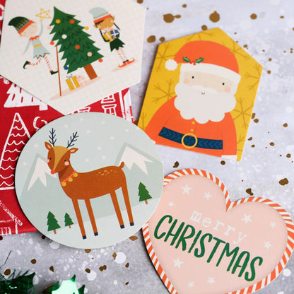 Four festive cards displayed on a glittery surface: one showcases elves decorating a tree with Printable Heaven’s Naughty or Nice Character Toppers (DCTOP208X21), another features Santa, one depicts a reindeer in the snow, and a heart-shaped card reads “merry CHRISTMAS”—a delightful way to add Printable Heaven charm to your holiday card toppers collection.
