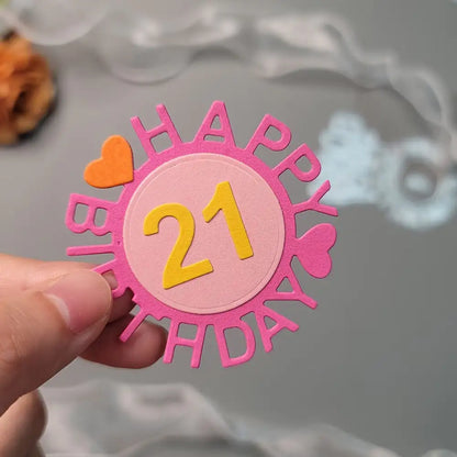 A hand holds a pink and yellow “Happy Birthday 21” cutout with heart shapes, crafted using the Printable Heaven Birthday Cutting Die Set with Numbers (4 PCS) (W092), against a blurred gray background.