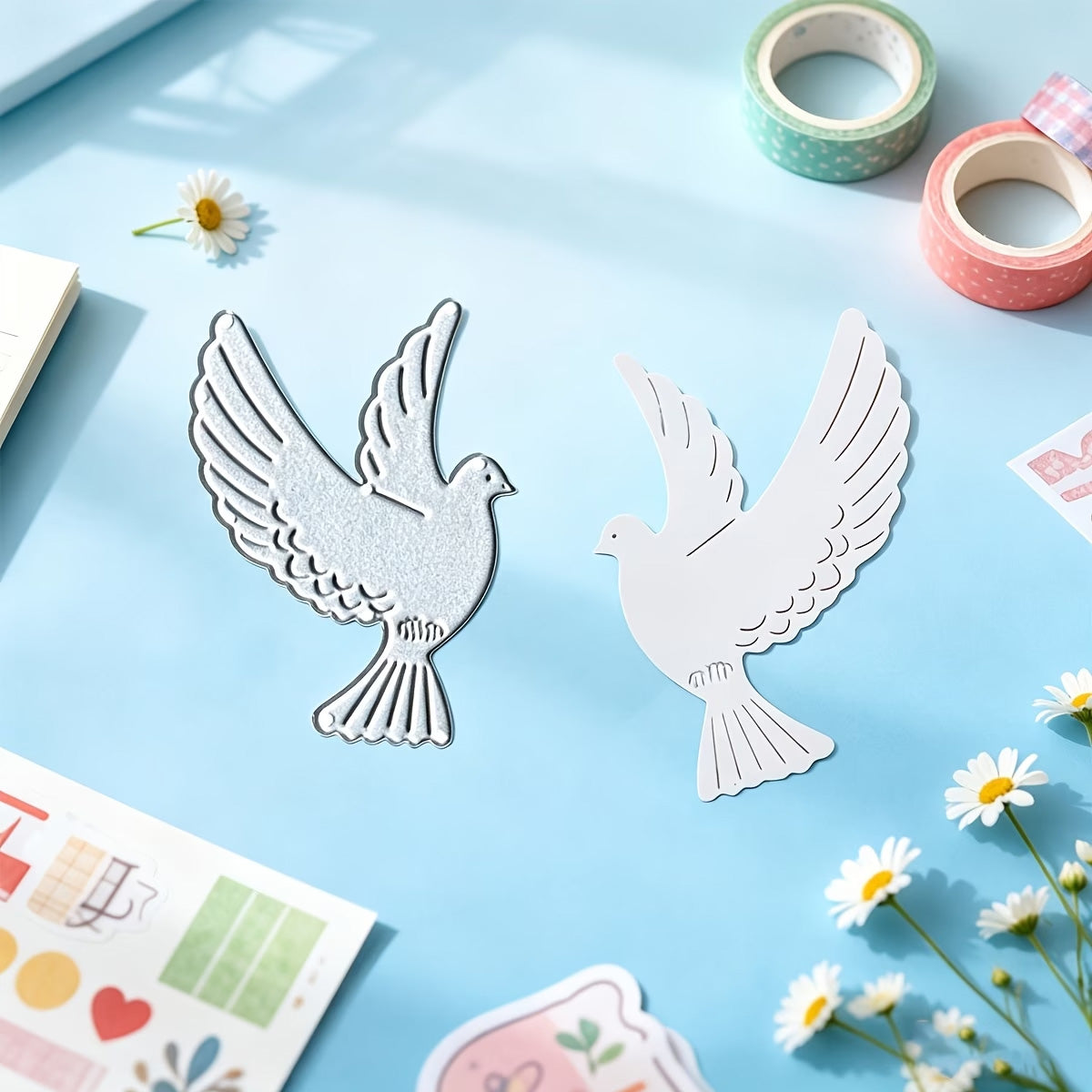 Two dove-shaped cutouts—one silver and one white—created using the Printable Heaven Flying Dove Metal Cutting Die (1 pc) (A020), are displayed on a light blue surface surrounded by daisies, colorful washi tapes, and stickers under bright lighting, offering cheerful card making inspiration.