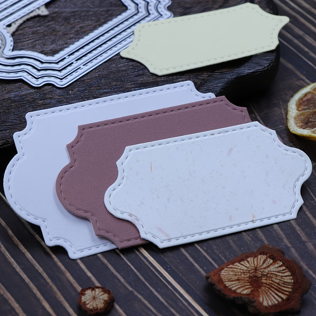 Printable Heaven’s Stitched Edge Nesting Frame Dies – Set of 4 (D124) are shown on a wooden surface beside dried fruit slices, along with assorted blank, decorative paper and metal labels featuring stitched edges—ideal for card making or pairing with nesting cutting dies.