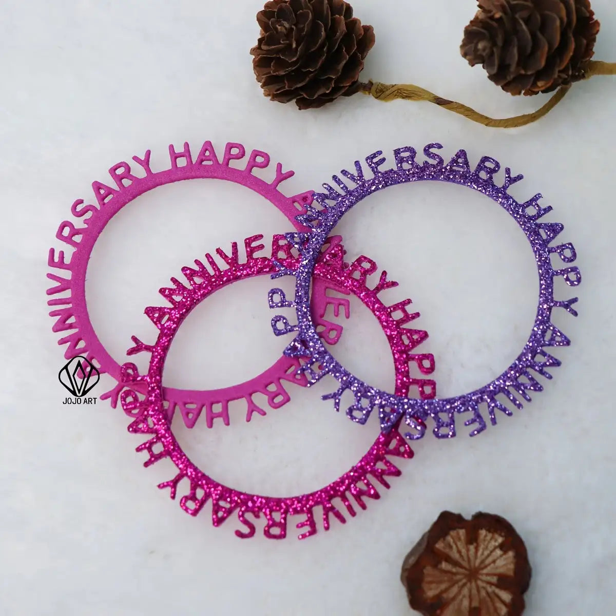 Four glittery “Happy Anniversary” die-cuts, created with the Printable Heaven Happy Anniversary Cutting Die (1 PC) (W151), two in pink and two in purple, are arranged overlapping each other on a white surface. Pine cones and a dried brown flower are placed nearby.