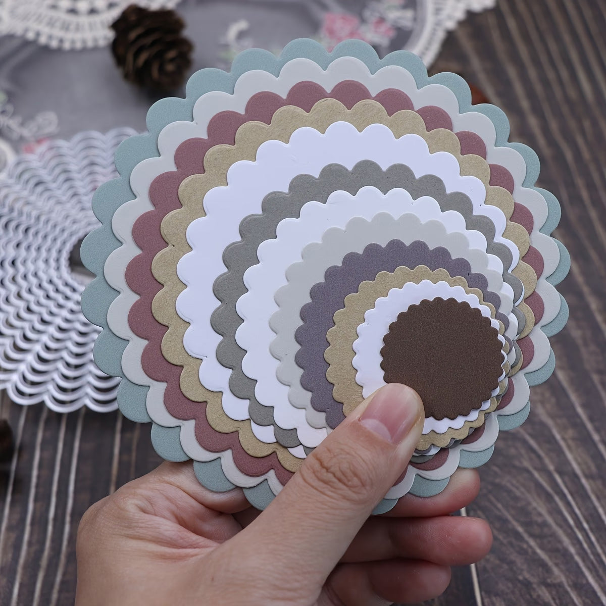 A hand holds a stack of scalloped, round paper cutouts—made using the Printable Heaven 12-Piece Scalloped Circle Cutting Die Set (D140)—in various sizes and colors such as white, brown, green, and peach, displayed on a wooden table with scrapbooking and card making supplies in the background.