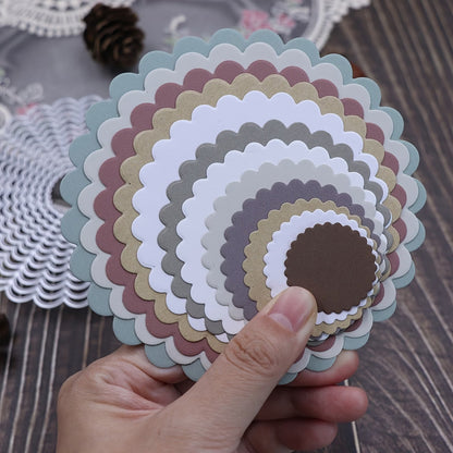 A hand holds a stack of scalloped, round paper cutouts—made using the Printable Heaven 12-Piece Scalloped Circle Cutting Die Set (D140)—in various sizes and colors such as white, brown, green, and peach, displayed on a wooden table with scrapbooking and card making supplies in the background.